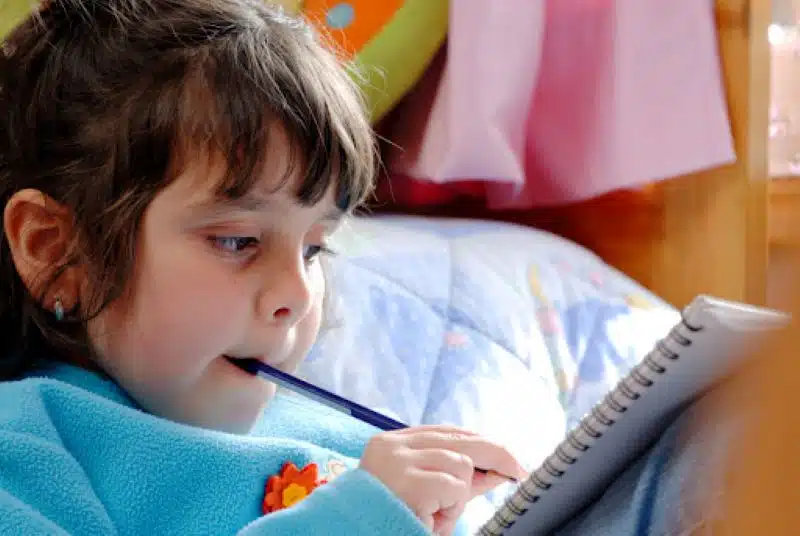 oung girl chewing on a pen while writing in a notebook, showcasing oral sensory seeking behavior in children.