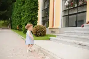 A toddler in a light blue dress stands at the foot of a stone staircase, looking up toward a building surrounded by green hedges.